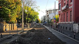 O zonă de șantier cu pământ întins este delimitată de garduri metalice pe o stradă cu clădiri și copaci pe margini.