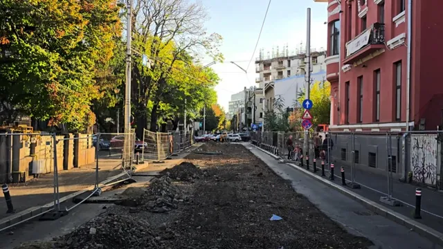 O zonă de șantier cu pământ întins este delimitată de garduri metalice pe o stradă cu clădiri și copaci pe margini.