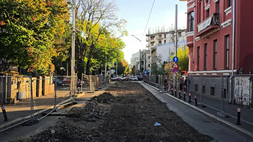 O zonă de șantier cu pământ întins este delimitată de garduri metalice pe o stradă cu clădiri și copaci pe margini.