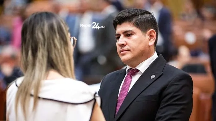 Un bărbat cu părul scurt și negru, purtând costum întunecat și cravată roz, privește spre o femeie cu părul blond în prim-plan.