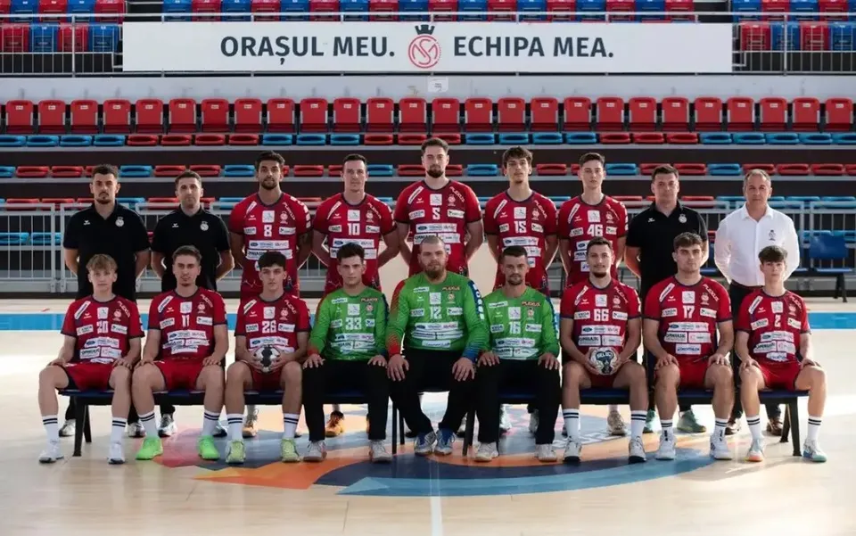 O echipă de handbal masculini poartă echipamente de sport roșii și verzi, fiind așezată în două rânduri pe un teren de sport interior.