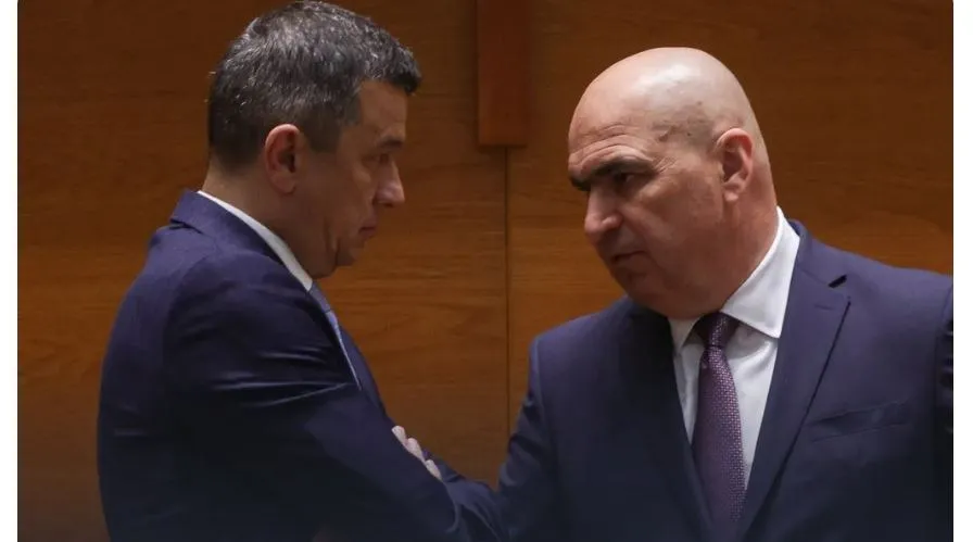 Doi bărbați în costume oficiale stau față în față într-o încăpere cu panouri de lemn în fundal.