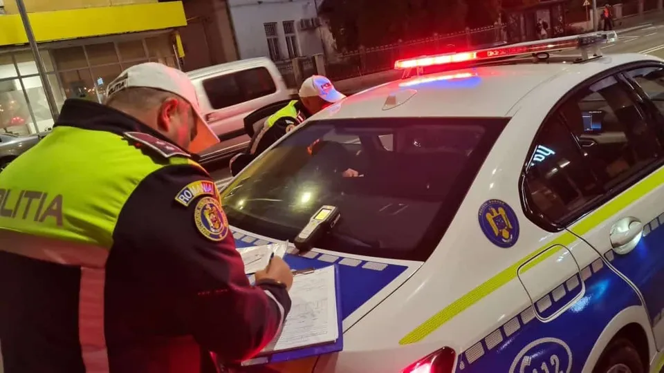 Un polițist în uniformă scrisă cu litere galbene notează pe o foaie lângă capota unei mașini de poliție cu girofaruri aprinse.