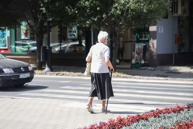 O femeie cu părul alb, purtând o bluză albă și o fustă neagră, traversează strada folosind un baston.