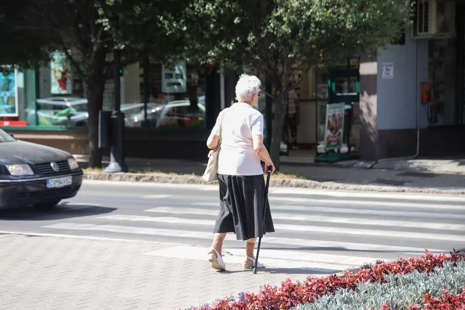 O femeie cu părul alb, purtând o bluză albă și o fustă neagră, traversează strada folosind un baston.