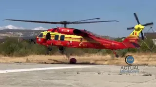 Un elicopter roșu și galben, posibil un Black Hawk, este fotografiat în aer, deasupra unei suprafețe de beton, cu vegetație uscată în fundal.
