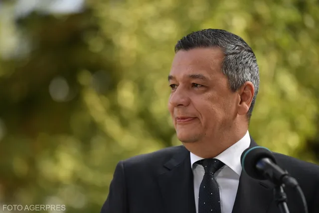 Un bărbat cu părul scurt și cărunt, purtând sacou negru, cămașă albă și cravată neagră, stă în fața unui microfon cu fundal verde neclar.