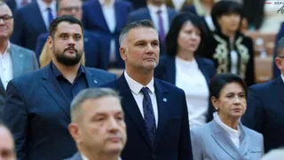 Un grup de persoane îmbrăcate în costume și haine formale stau în picioare într-o încăpere cu scaune de tip auditorium.