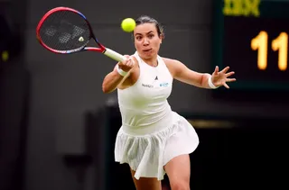 O femeie cu haine albe ține o raketă de tenis și privește o minge galbenă în aer pe un teren de sport.