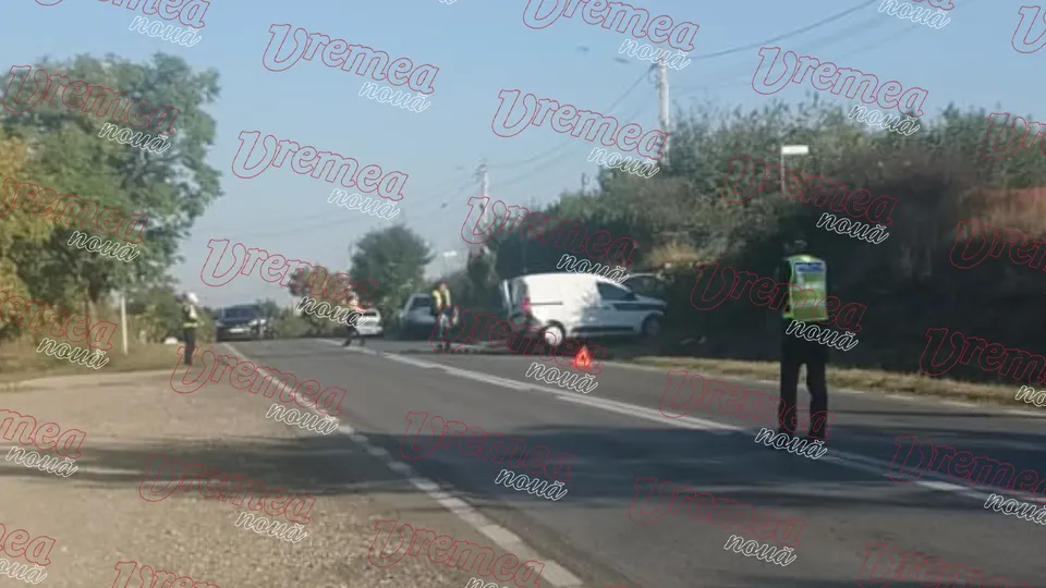 O persoană cu vestă reflectorizantă stă pe marginea unui drum asfaltat lângă un vehicul alb și un con de circulație portocaliu.
