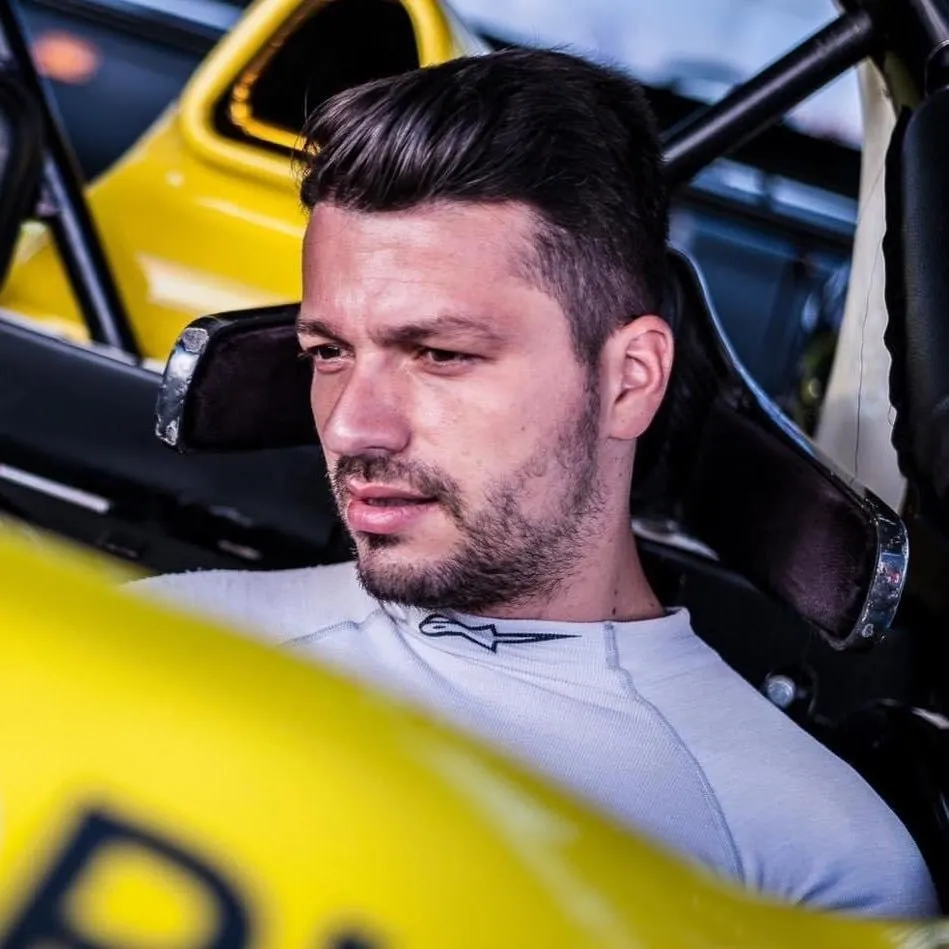 Un bărbat cu barbă scurtă și tricou alb stă într-un vehicul de curse galben cu structură de protecție neagră.