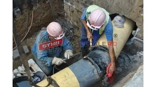 Două persoane purtând căști de protecție și mănuși lucrează la o conductă mare aflată într-o șanț de excavare.