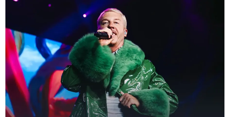 Un bărbat cu părul scurt și deschis la culoare poartă o haină verde lucioasă cu guler de blană și cântă într-un microfon.