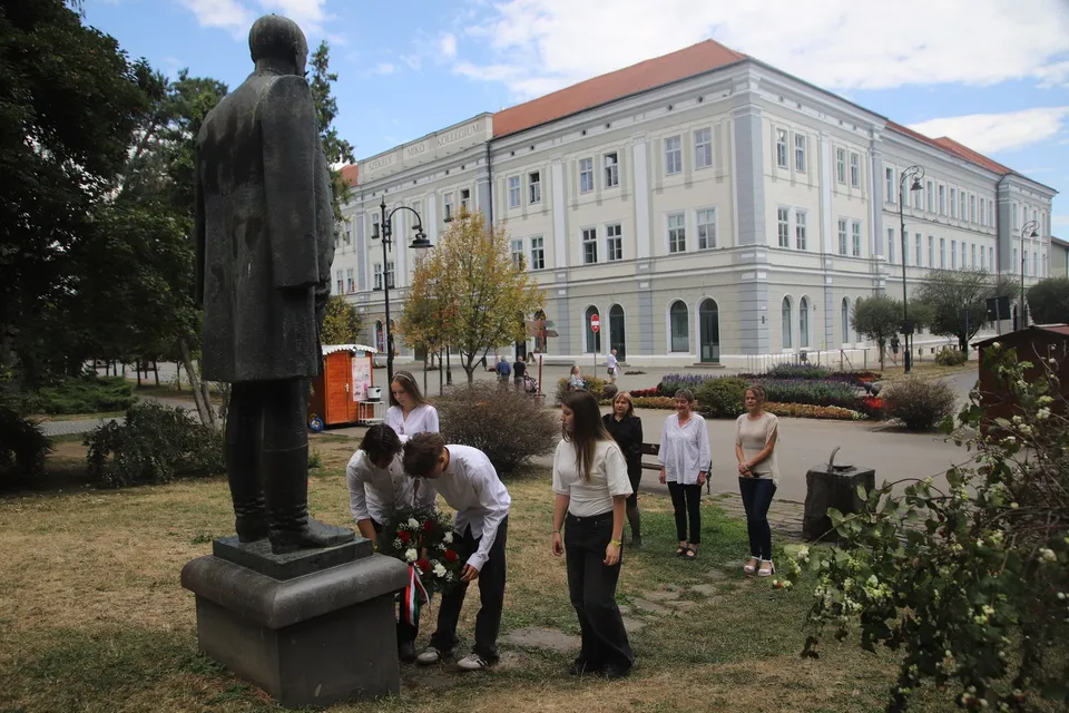 Un grup de persoane stă pe un teren cu iarbă lângă o statuie și în fața unei clădiri mari de culoare deschisă.