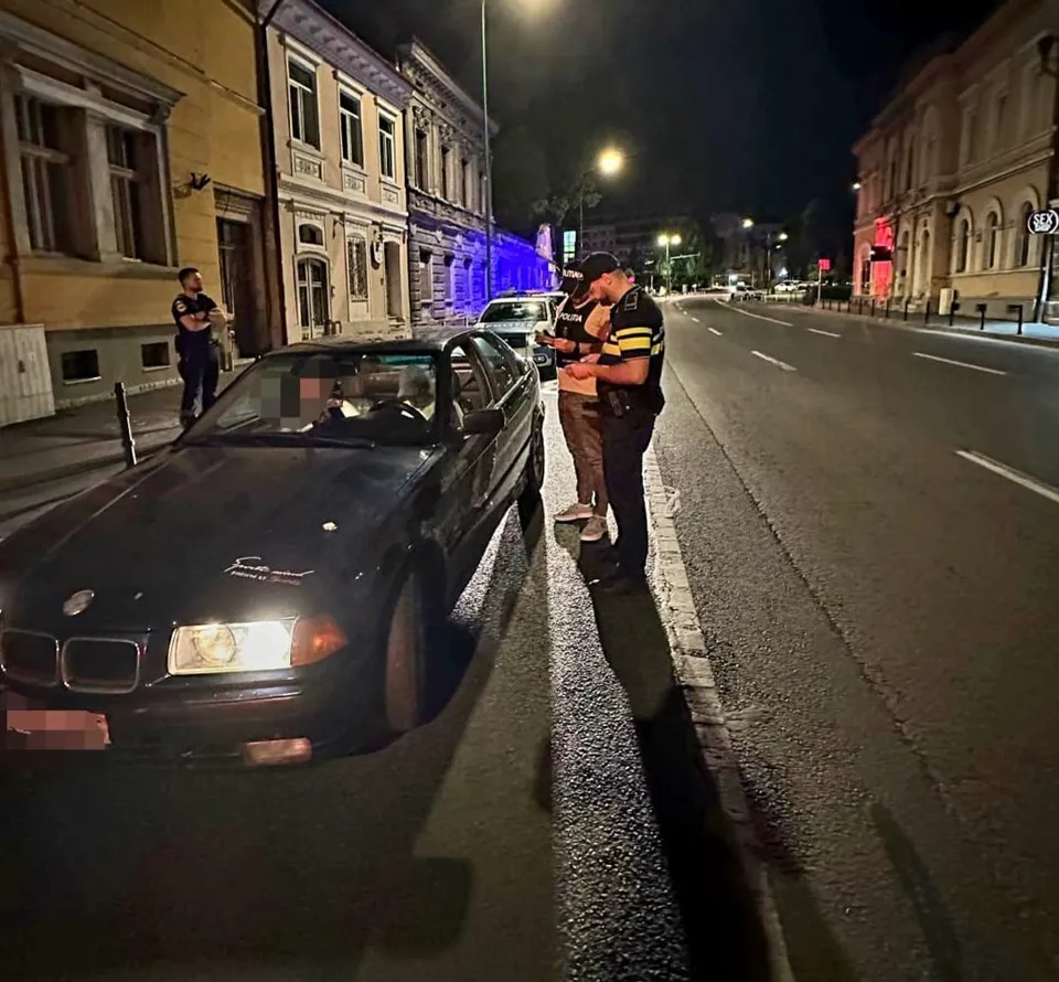 Unii polițiști în uniformă stau lângă o mașină neagră parcată pe marginea unui drum urban iluminat noaptea.