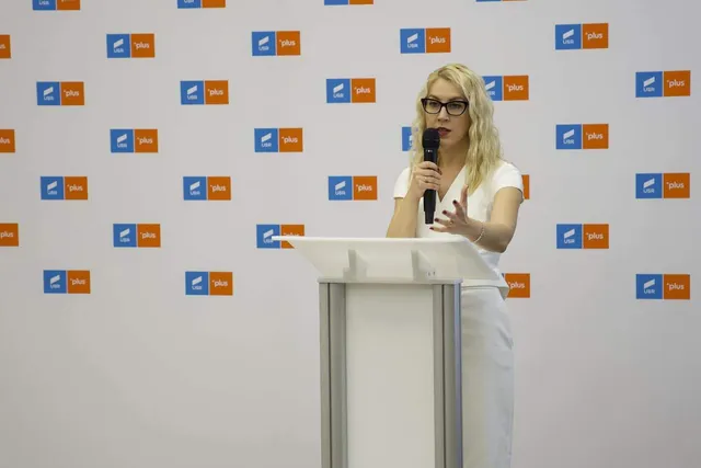 O femeie cu părul blond și ochelari stă în spatele unui podium alb, ținând un microfon în mână în fața unui fundal cu logo-uri repetate.