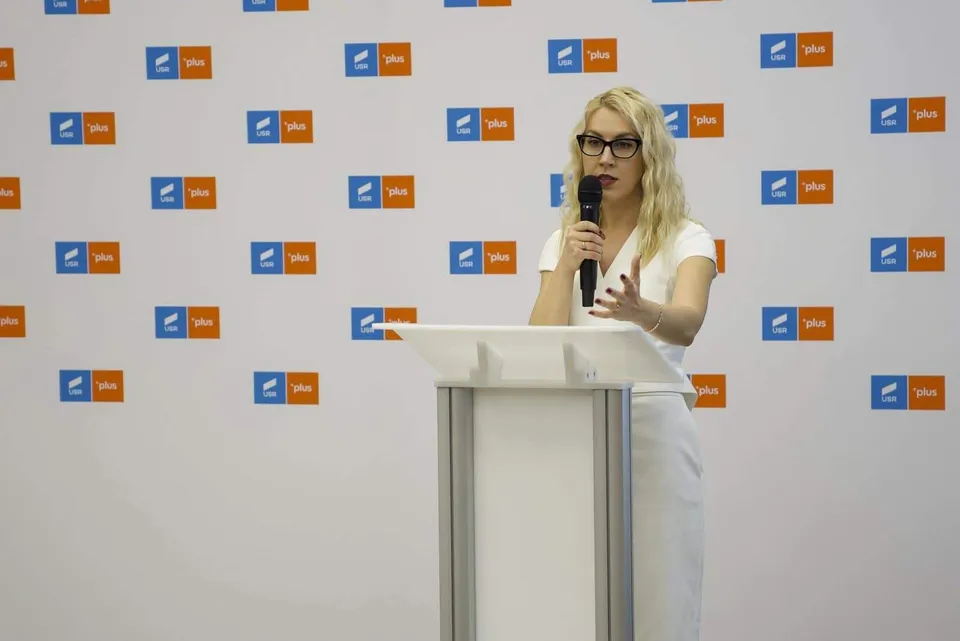 O femeie cu părul blond și ochelari stă în spatele unui podium alb, ținând un microfon în mână în fața unui fundal cu logo-uri repetate.