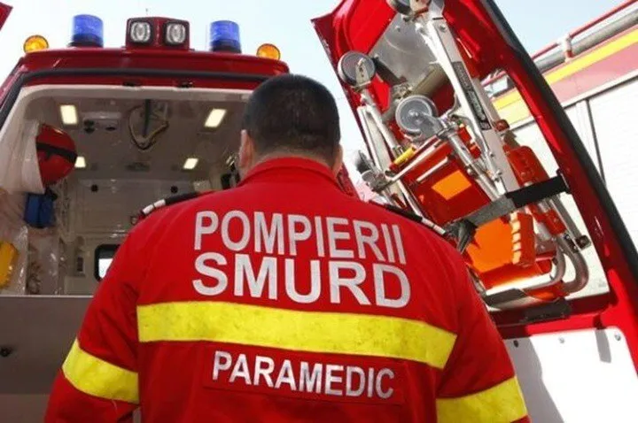 Un membru al echipei de intervenție cu uniformă roșie și benzi galbene pe spate stă lângă un vehicul de serviciu deschis.