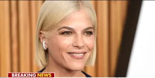 O femeie cu părul blond și cercei argintii zâmbește în fața unui fundal maro cu linii verticale.