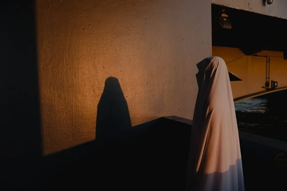 O persoană acoperită de un văl alb stă în fața unui perete sub lumina unei surse de iluminat.