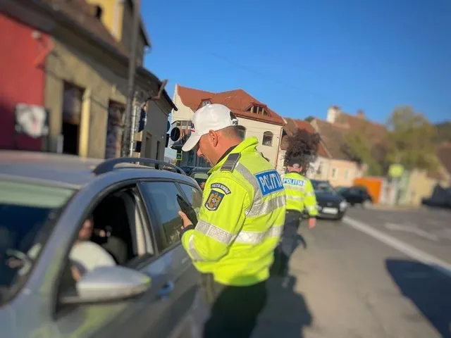 Un polițist cu vestă reflectorizantă galbenă stă lângă o mașină gri pe un drum urban lângă clădiri sub un cer senin.