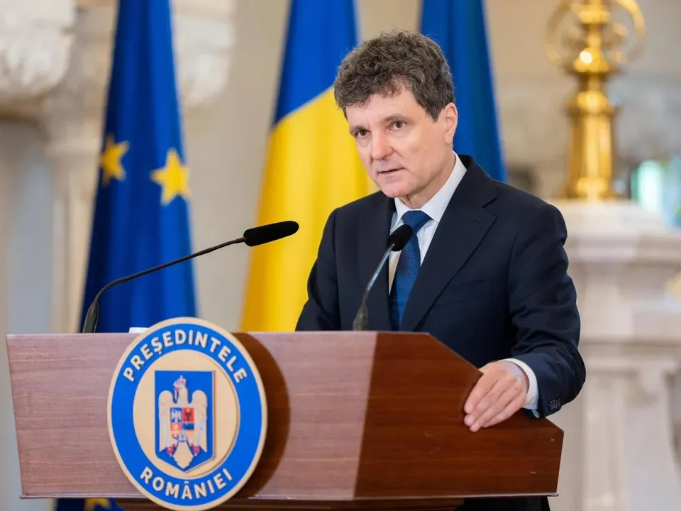 Un bărbat cu părul scurt și cămașă albă, purtând sacou închis și cravată albastră, stă în spatele unui podium de lemn cu un emblema oficială.