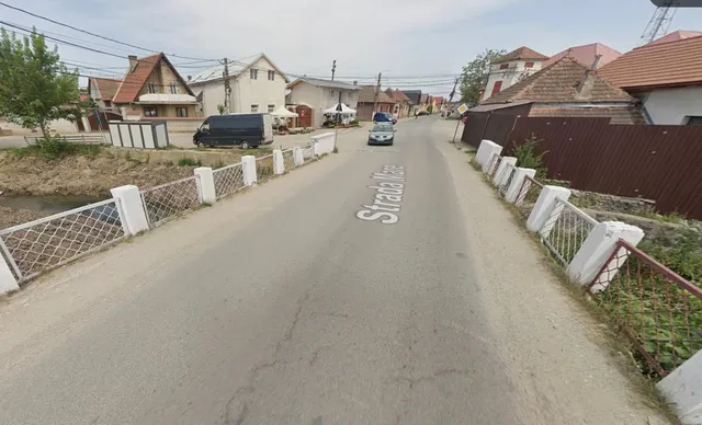 O stradă asfaltată cu marcaj alb trece peste un pod delimitat de balustrade albe și metalice, având case și vehicule în fundal.