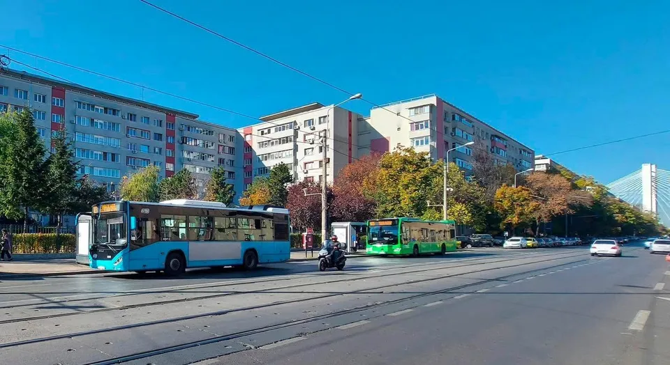 Un autobuz albastru și unul verzi circulă pe o stradă cu șine de tramvai, lângă blocuri de apartamente sub un cer senin.