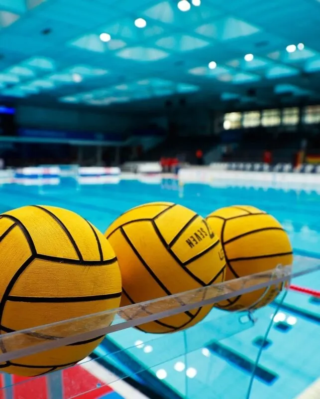 Trei mingi galbene cu linii negre sunt așezate pe un marginea transparentă a unei piscine interioare.