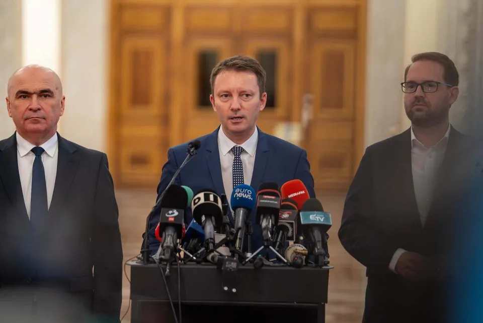 Trei bărbați în costume stau în spatele unui podium pe care sunt așezate numeroase microfoane de presă.