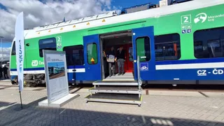 Un tren verde și albastru cu ușa deschisă este parcat lângă niște trepte metalice pe un peron de pavaj sub un cer înnorat.