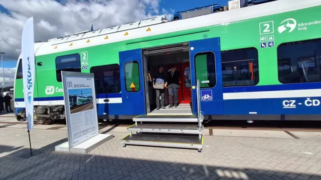 Un tren verde și albastru cu ușa deschisă este parcat lângă niște trepte metalice pe un peron de pavaj sub un cer înnorat.