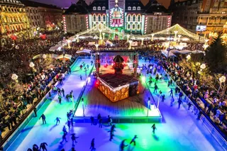 O mulțime de oameni se adună în jurul unei piste de patinaj iluminată colorat, aflată într-o piață publică cu clădiri decorate noaptea.