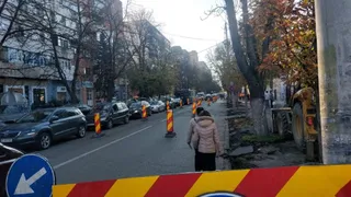 Mașini sunt parcate pe marginea unui drum asfaltat lângă clădiri înalte, în timp ce un om stă pe trotuar lângă un utilaj galben.