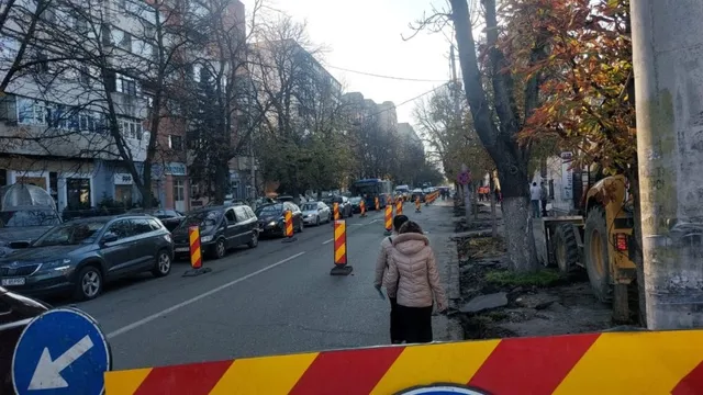 Mașini sunt parcate pe marginea unui drum asfaltat lângă clădiri înalte, în timp ce un om stă pe trotuar lângă un utilaj galben.