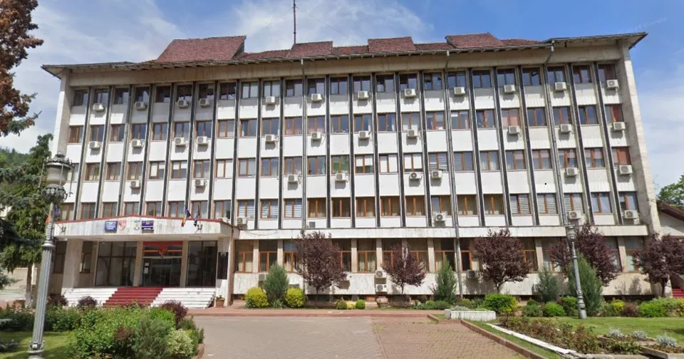 O clădire administrativă cu mai multe etaje, ferestre multiple și unități de aer condiționat, situată în spatele unui spațiu verde cu arbuști.