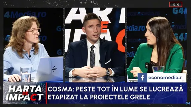 Trei persoane stau la o masă într-un studio de televiziune, în timp ce un bărbat în costum și cravată vorbește în centru.