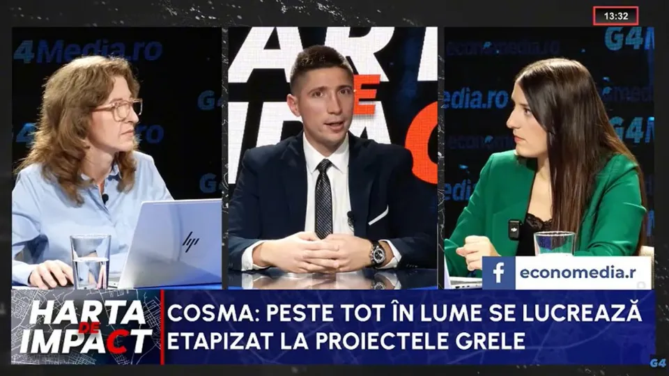 Trei persoane stau la o masă într-un studio de televiziune, în timp ce un bărbat în costum și cravată vorbește în centru.