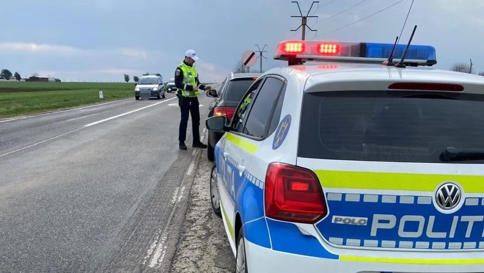 Un polițist cu vestă reflectorizantă stă lângă un vehicul roșu pe marginea unui drum, lângă o mașină de poliție albastră și albă.