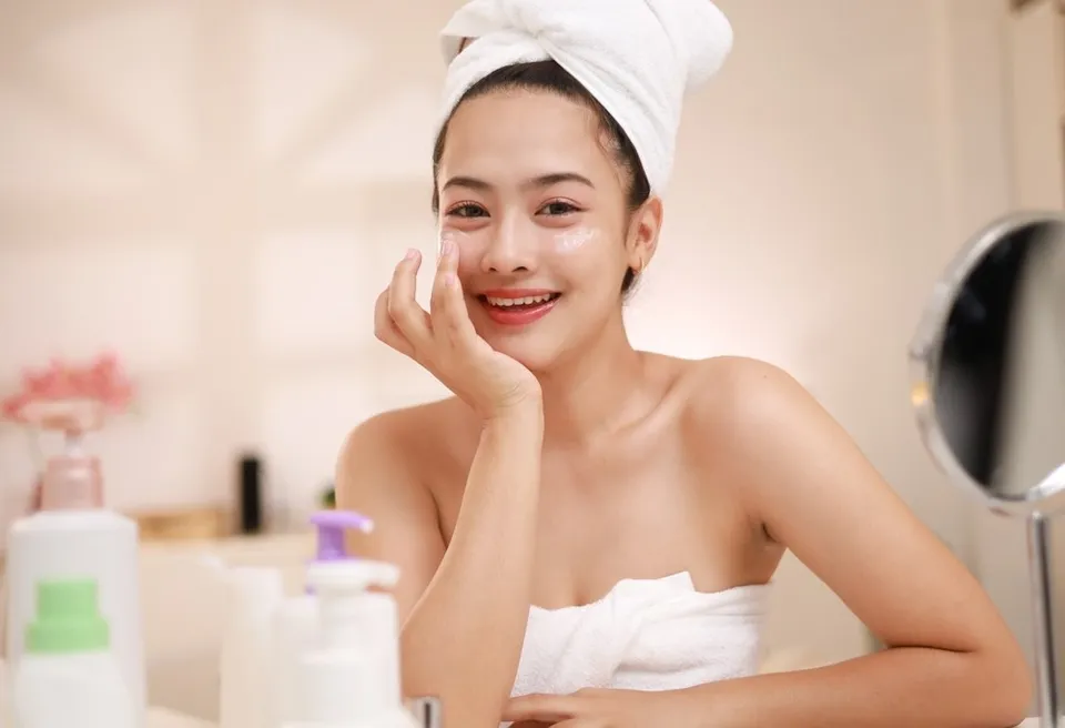 O femeie cu prosop alb pe cap aplică o cremă albă pe obrazul drept, stând în fața unei oglinzi lângă diverse flacoane de produse cosmetice.