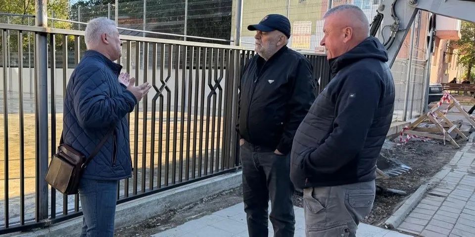 Trei bărbați stau pe un trotuar lângă un gard metalic negru, unul purtând o geantă de umăr și o jachetă albastră.