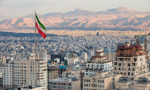 Steagul iranian flutură pe un stâlp deasupra unui peisaj urban dens cu clădiri înalte și munți în fundal.