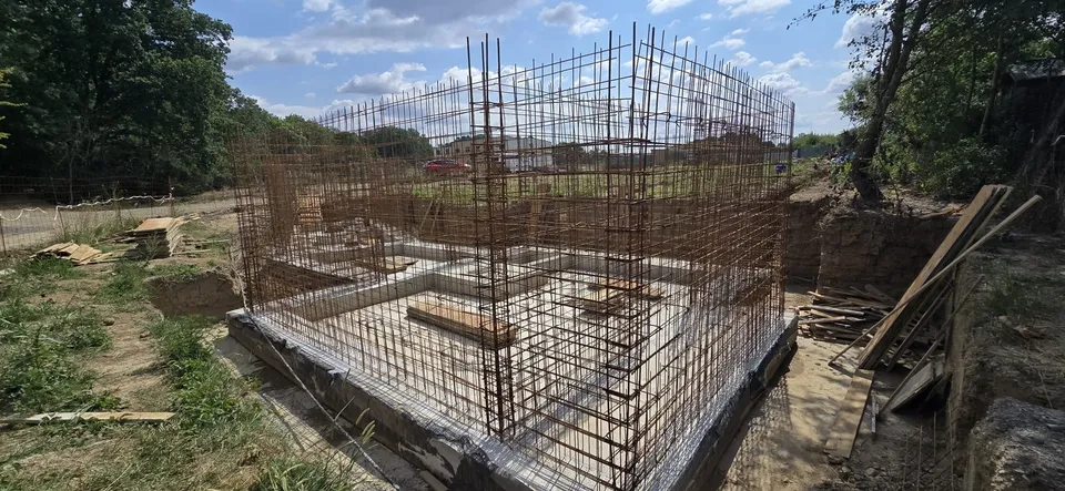 O structură de armătură metalică este instalată pe o fundație de beton într-un șantier de construcții aflat în aer liber.