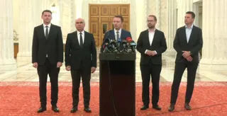 Cinci bărbați în costume scurte stau în picioare pe un covor roșu în fața unui podium cu microfoane.