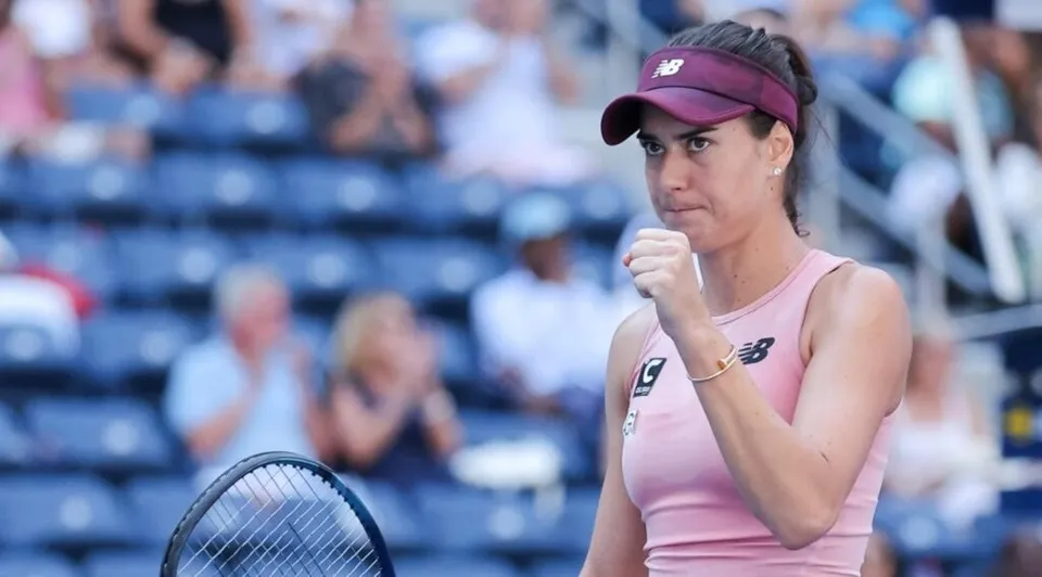 Fotografie de arhivă cu o jucătoare de tenis în roz, purtând o căciulă mov și ținând o raclă pe un teren cu tribune.