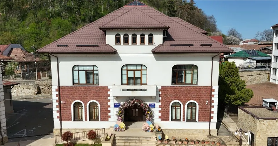 O clădire albă cu acoperiș maro și detalii din cărămidă roșie prezintă ferestre arcuate și un aranjament floral deasupra intrării principale.
