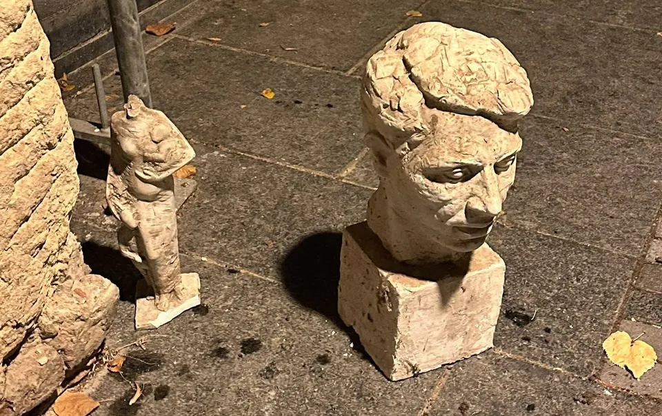 Două sculpturi din piatră, un bust și o figură umană fragmentată, sunt așezate pe un podea de pavaj întunecată.