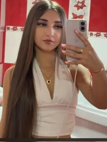 O tânără cu părul lung și deschis la culoare poartă un top alb și un colier cu pandantiv, făcând un selfie în oglindă.