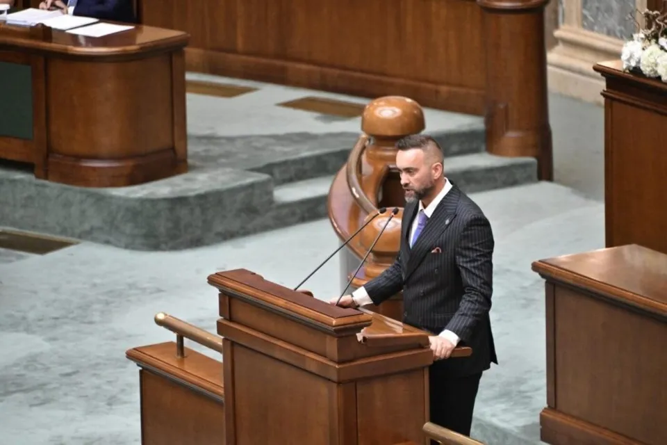 Un bărbat cu barbă, îmbrăcat într-un costum în dungi, stă în picioare în fața unui pupitru de lemn într-o încăpere cu mobilier din lemn.