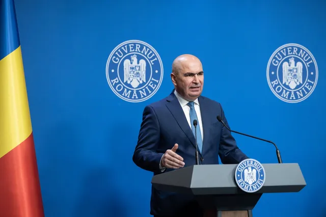 Un bărbat cu costum închis și cravată stă în spatele unui podium cu microfoane în fața unui fundal albastru cu sigilii oficiale.